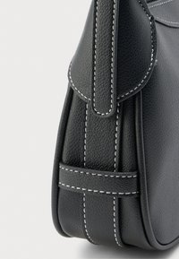 Coin d'un sac à main en cuir noir texturé avec couture blanche et détail de sangle pliée sur un fond clair uni.