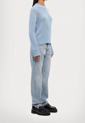 Vrouw staand opzij, gekleed in een lichtblauwe gebreide trui, lichtgewassen spijkerbroek en zwarte chunky platform schoenen, tegen een witte achtergrond.