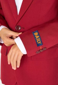 Blazer rosso con finitura testurizzata, dotato di chiusura a bottone e di un'etichetta blu ricamata con la scritta “DAILY” sul polsino.
