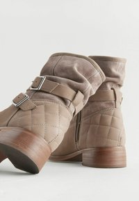 Bottines beiges avec un design matelassé, en suède, accents à double sangle et un petit talon en bois. Fermeture éclair sur le côté.
