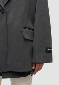 Blazer in lana grigia scura con tasca a patta, maniche lunghe con una toppa nera contenente testo bianco, indossato su pantaloni crema che mostrano una mano e le dita.