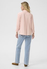 Camicia a maniche lunghe rosa chiaro con vestibilità rilassata, caratterizzata da maniche arricciate. Abbinata a jeans blu chiaro e scarpe marroni slip-on.
