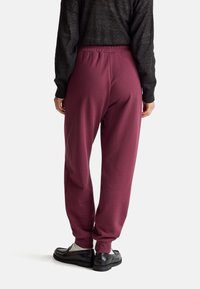 Bordeauxrote Sweatpants mit einem elastischen Bund und schmal zulaufenden Beinen, kombiniert mit schwarzen Slippern. Das Material scheint weich und leicht zu sein.
