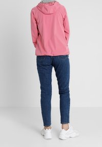 Rosa lättviktsjacka med huva, grå foder och elastiska ärmslut. Bärs med blå jeans och vita sneakers. Enkel bakgrund.