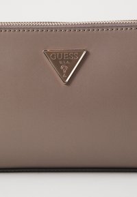 Bruine neplederen portemonnee met ritssluiting. Bevat een driehoekig metallic logo met "GUESS" en "U.S.A." in reliëf. Soepele textuur.