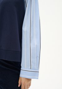 Top stratificato con un corpo blu navy e maniche a strisce blu chiaro con accenti neri e bianchi. Il materiale è morbido con una consistenza evidente.