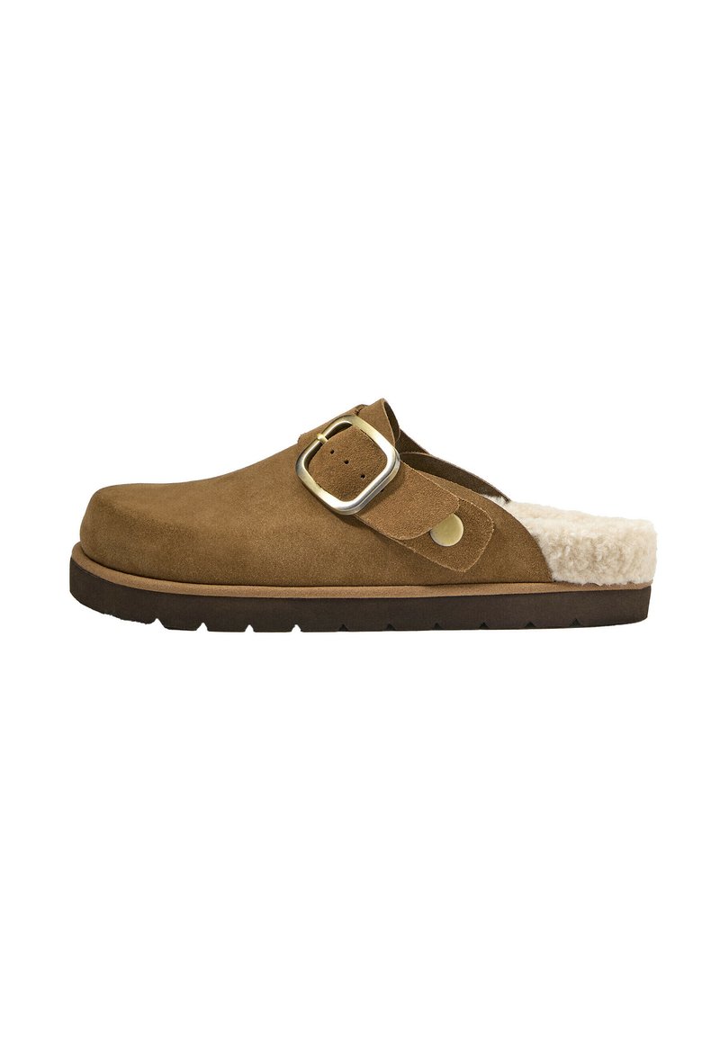 Clog din suede maro, slip-on, cu cataramă aurie și căptușeală din blană crem, talpă plată de culoare închisă, profil lateral pe fundal alb.