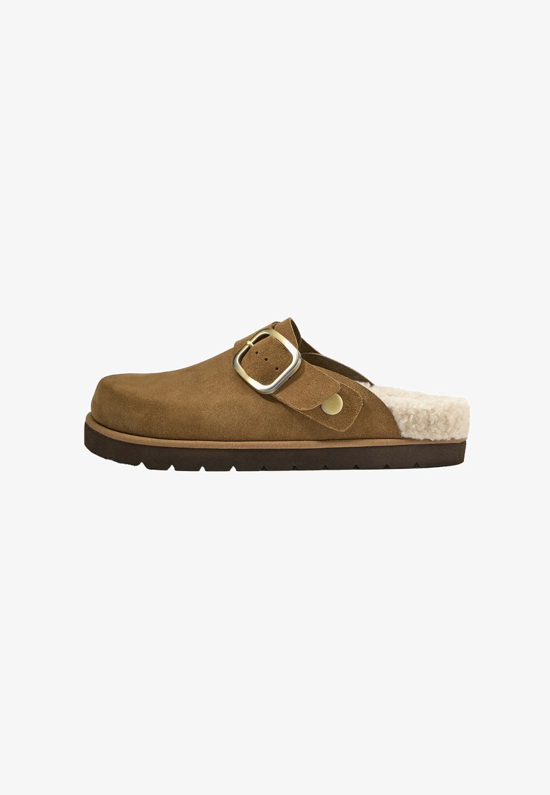 Clog din suede maro, slip-on, cu cataramă aurie și căptușeală din blană crem, talpă plată de culoare închisă, profil lateral pe fundal alb.