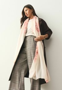 Viegls, rakstains šalle maigi rozā un krēmkrāsā, kārtās pār baltu topu un pelēkas rūtainas plašas bikses, kopā ar tumšu mēteli.