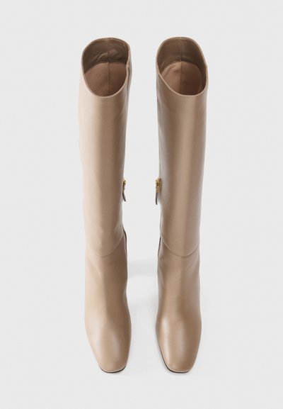Knee-high beige leather boots with a smooth finish, square toe, and gold zipper accents on the outer side. Flat sole design.