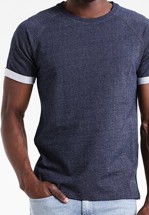 Man draagt een donkerblauw T-shirt met korte mouwen en een ronde hals, met witte mouwboorden, hand in de zak en gecombineerd met lichtblauwe spijkerbroek.