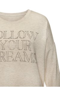 Béžová dlhá rukávová košeľa s širokým výstrihom a jemnou textúrou. Na prednej strane je nápis "FOLLOW YOUR DREAMS" vyrobený z kamienkov.