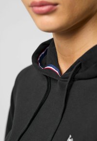 Zwarte hoodie met een gestructureerd oppervlak, voorzien van een meerkleurige rand aan de binnenkant van de hals en een klein logo aan de voorkant.