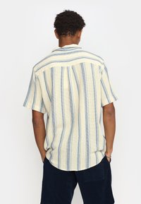 Camisa de manga corta y cuello en tela color crema con rayas verticales azules. Presenta un dobladillo recto y un ajuste relajado. Material texturizado.
