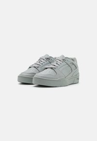 Un par de zapatillas Puma de color gris claro con cordones, que presentan paneles texturizados y punteras perforadas, mostradas desde un ángulo frontal sobre un fondo blanco.