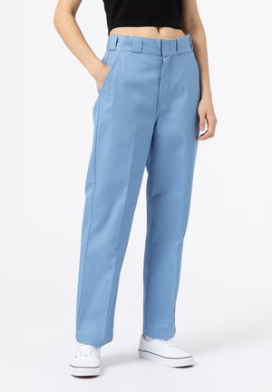 Person iført lyseblå bukser med vide ben, sort crop top og hvide sneakers, med den ene hånd i lommen, stående mod en hvid baggrund.