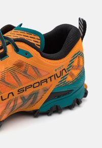 Παπούτσι trail με ένα πορτοκαλί και θαλασσί επάνω μέρος, που διαθέτει υφή και μαύρες λεπτομέρειες, σύνθετη σόλα με πλίνθους για πρόσφυση.