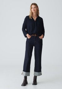 Camicia blu marina con bottoni, pantaloni larghi in denim scuro con risvolti; cintura marrone, stivali alla caviglia marroni, texture lisce.