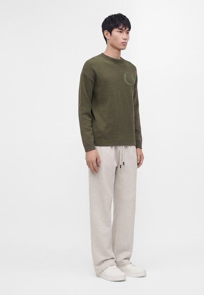 Homme portant un pull vert olive, un pantalon beige à cordon et des baskets blanches, debout devant un fond blanc uni.