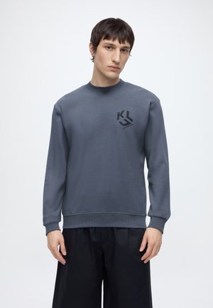 Ung mand med kort mørkt hår iført en blå-grå sweatshirt med sort logo på brystet og løse sorte bukser, stående mod hvid baggrund.