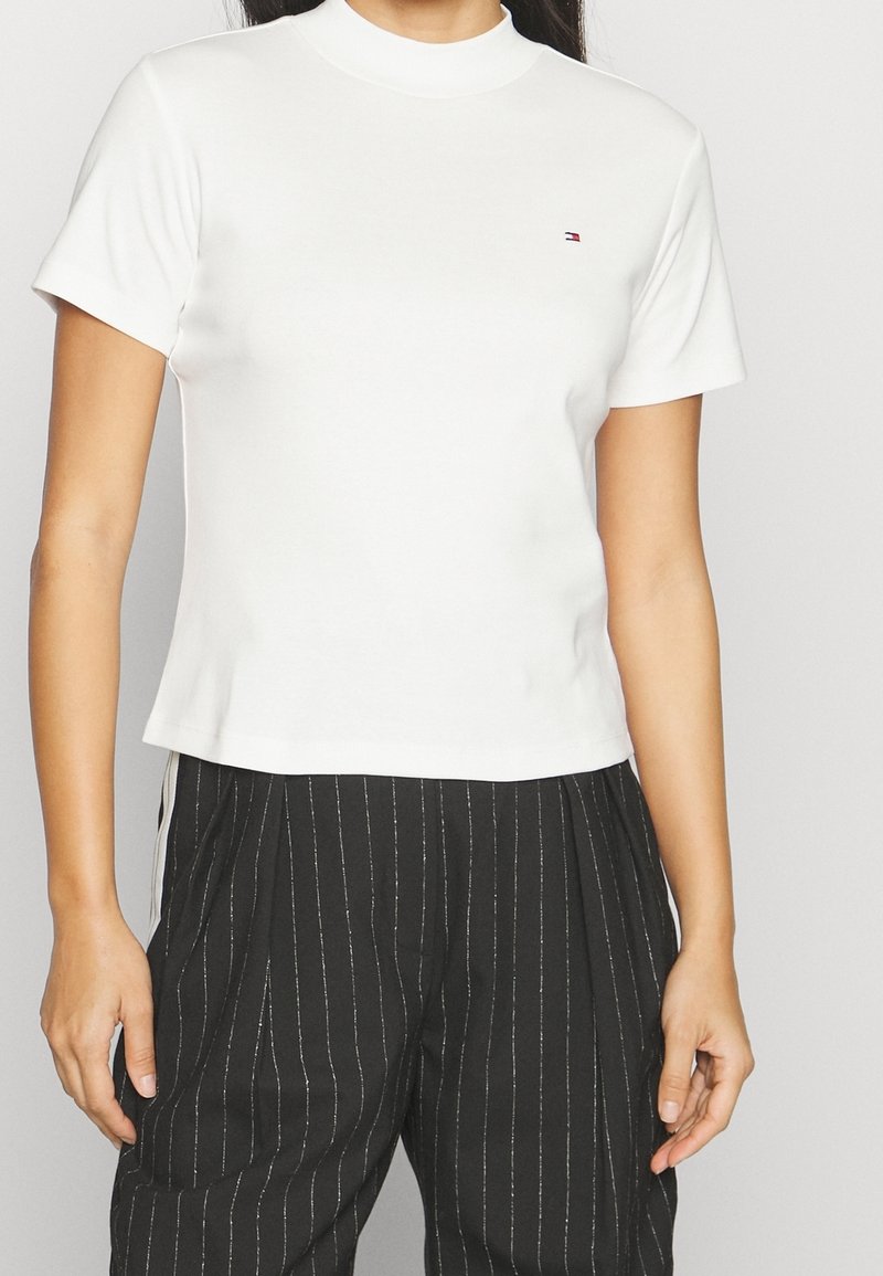 Bílé tričko s krátkým rukávem a vysokým límečkem s přiléhavým střihem a malým vyšívaným logem na hrudi, doplněné o černé kalhoty s proužky.