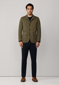 Giacca leggera verde oliva con due tasche frontali, una fila di bottoni e colletto a punta, abbinata a pantaloni scuri e scarpe marroni.