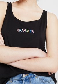 Mustat hihaton paita, jossa on alhaalla oleva kaula-aukko ja monivärinen "WRANGLER" -logo. Kangas näyttää pehmeältä; yhdistetty vaaleansinisiin farkkushortsien kanssa.