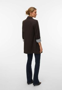 Smeđi dugi kaput s ovratnikom, ¾ rukavima sa sivim prugastim manšetama i ravnim krojem. Nošen preko tamnih zvonolikih traperica.