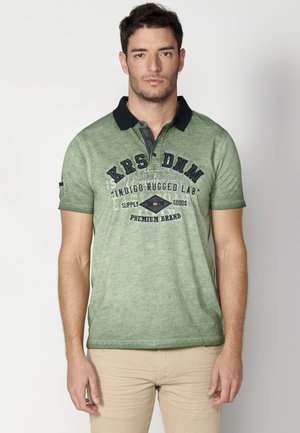 Polo vert avec un col sombre, à manches courtes, et un imprimé graphique sur le devant. Le tissu semble texturé avec un subtil motif chiné.