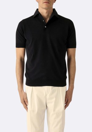 Uomo che indossa una maglia nera a maniche corte con colletto a maglia e pantaloni beige plissettati su sfondo neutro.