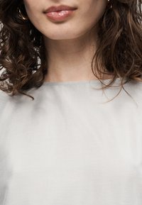 Femme aux cheveux bruns bouclés portant un haut gris clair, affichant des lèvres avec un rouge à lèvres rose brillant ainsi qu'une partie de son cou et de ses épaules.