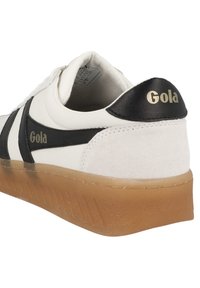 Sneaker bianca con accenti neri e tallone texturizzato. La suola in gomma è traslucida e presenta un design con battistrada. Marchio in oro.