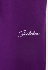 Fioletowy materiał bluzy z haftowanym napisem "Smélodox Essentials" w białym i zielonym kolorze. Gładka tekstura bez widocznych wzorów.