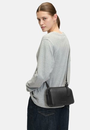 Schwarze Ledertasche im Querformat mit Reißverschluss, rechteckiger Form und glatter Textur, getragen auf einem grauen Langarmtop.