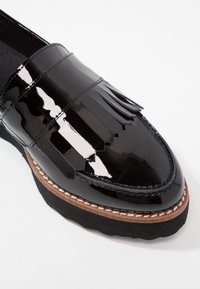 Mocassin en cuir vernis noir avec détail de franges, bordure épaisse marron, coutures blanches et semelle crêpe noire sur fond blanc.