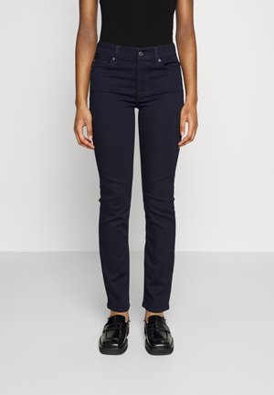 Persoon draagt aansluitende donkerblauwe broek, zwart topje en glanzende zwarte loafers, staand tegen een effen lichte achtergrond.