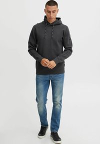 Mørkegrå hættejakke med snoretræk, tekstureret stof, båret over blå jeans, parret med sorte sneakers, stående på en hvid baggrund.