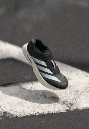 Black athletic shoe featuring three white stripes, textured upper, sleek design, and a curved sole, positioned above a white line on asphalt.