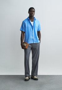 Άνδρας με φωτεινό μπλε πουκάμισο κοντομάνικο, γκρι παντελόνι και δίχρωμα παπούτσια, κρατώντας ένα μικρό καφέ βιβλίο σε απλό φόντο.