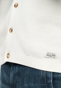 Približnji posnetek bele teksturirane jopice s rjavimi gumbi in etiketo "Guess Jeans 1981" nad temno modrimi denim kavbojkami.