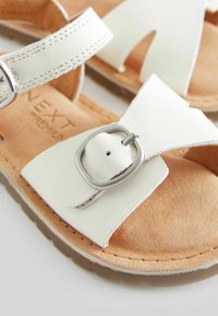 Vita syntetiska lädersandaler med en texturerad beige sula. Funktioner inkluderar ett bälte med en silverfärgad spänne och öppen tå-design.