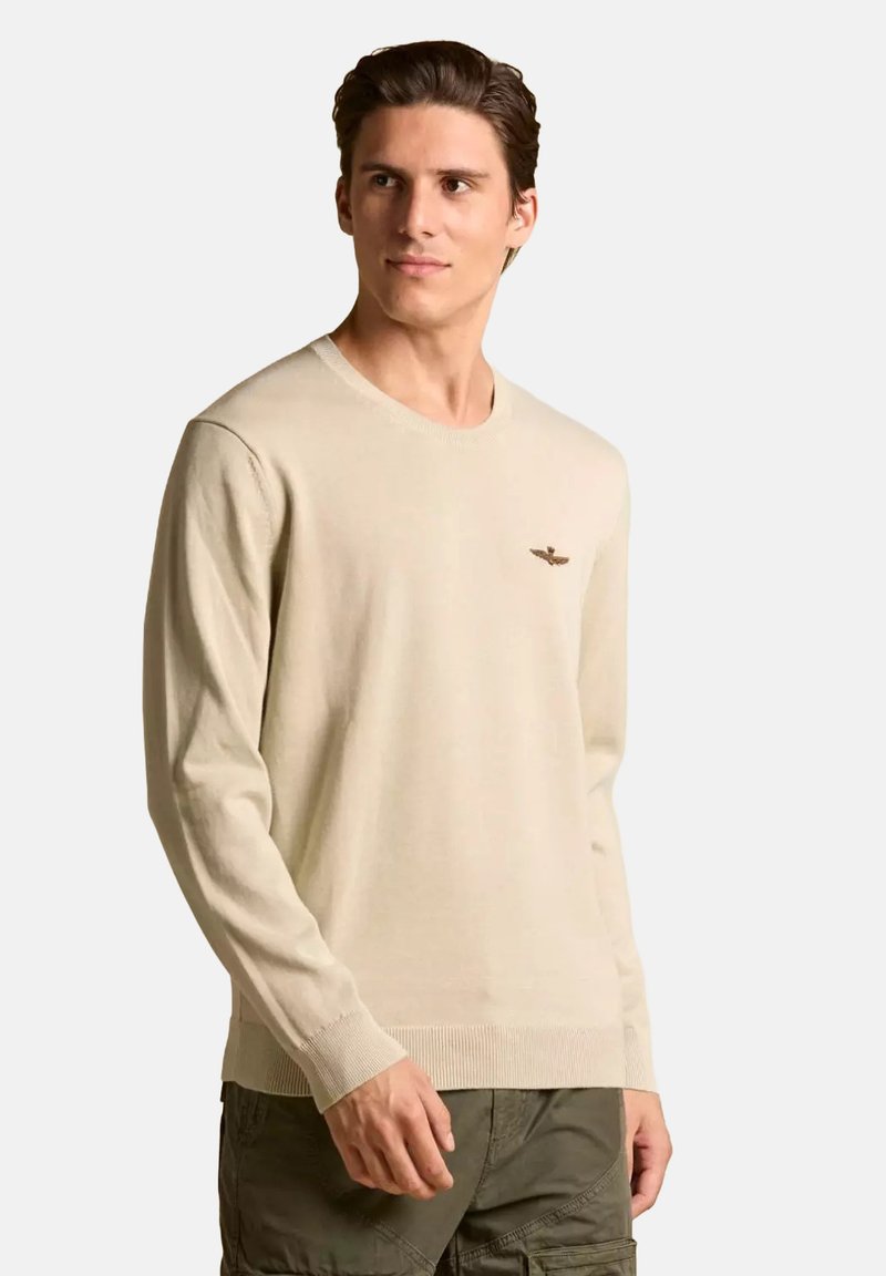 Maglione lavorato a maglia beige con maniche lunghe, scollo rotondo e polsini a coste. Presenta un piccolo logo ricamato sul lato sinistro del petto.