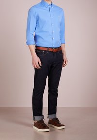 Světle modrá košile s dlouhými rukávy, tmavé džíny, hnědý kožený opasek a hnědé tenisky s bílou podešví. Neformální outfit.