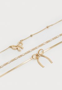 Drie verfijnde gouden kettingarmbanden, twee met strikvormige bedels en één met kleine ronde kralen op een effen witte achtergrond.