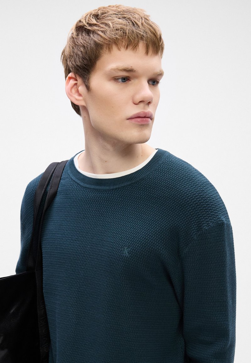 Teal textured sweater with a crew neck and a small emblem on the chest, paired with a dark shoulder bag. White inner collar visible.