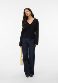 Svart cardigan med V-ringning och knappdetalj, kombinerad med mörkblå vida jeans. Modellen håller en gul väska; svarta pekade skor.
