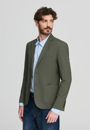 Blazer verde oliva con un design strutturato, stile monopetto, revers a punta e due tasche frontali, abbinato a una camicia azzurra.
