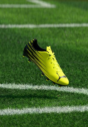 Żółty i czarny but piłkarski Puma unoszący się nad zielonym trawiastym boiskiem w pobliżu białych namalowanych linii.