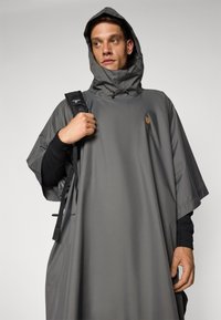 Poncho imperméable gris avec capuche, détails en cordon de serrage et logo, porté par-dessus une chemise noire à manches longues, avec une coupe décontractée.
