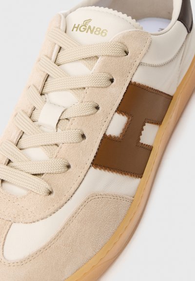 Zapatilla beige con una combinación de materiales de ante y cuero. Cuenta con un logo marrón prominente y cordones planos beige, con suela de goma.